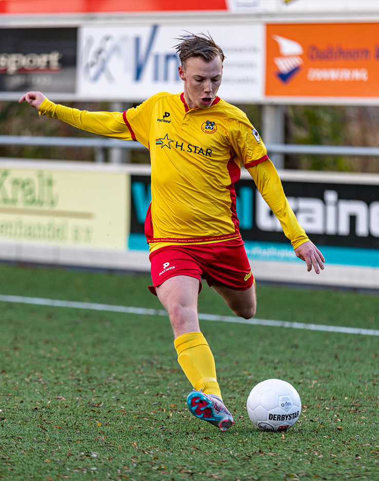 Voetballer in geel en rood tenue schiet de bal op het veld